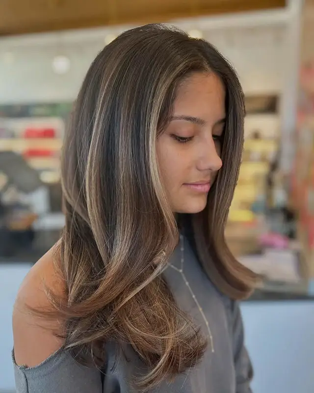 A young woman with long, layered brown hair with subtle highlights looks downward in a softly lit indoor setting. - Mango Salon, VA