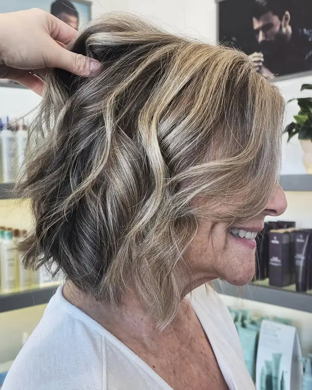 A woman with short, wavy, blonde and brown highlighted hair is shown in profile at a salon. A hand is lifting a section of her hair. - Mango Salon, VA