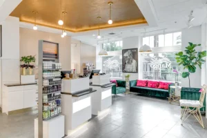 Modern spa reception area with product displays, green velvet seating, potted plants, large windows, and gold ceiling accents. - Mango Salon, VA