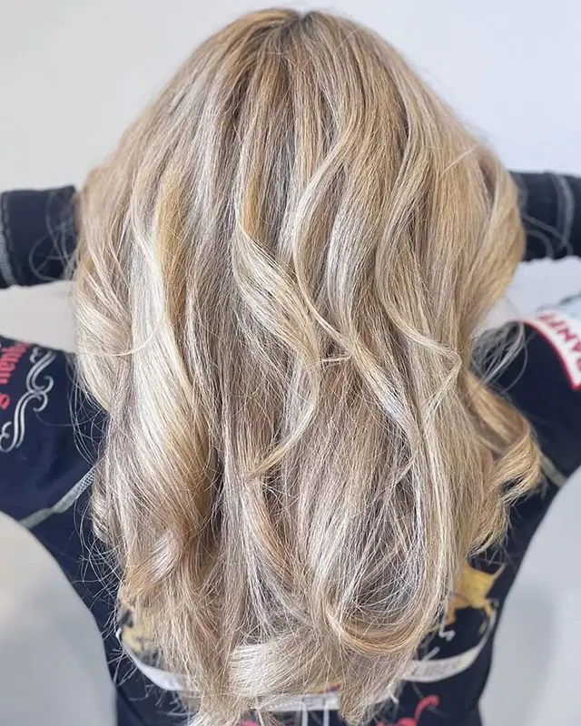 Person with long, wavy blonde hair photographed from behind, wearing a dark patterned shirt with arms raised and hands behind their head. - Mango Salon, VA
