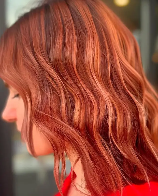 A person with wavy, shoulder-length copper red hair is shown in profile, wearing a red top. - Mango Salon, VA