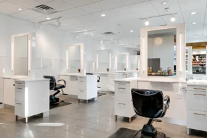 A modern hair salon interior featuring multiple styling stations with mirrors, black chairs, white countertops, and shelves of hair products in the background. - Mango Salon, VA