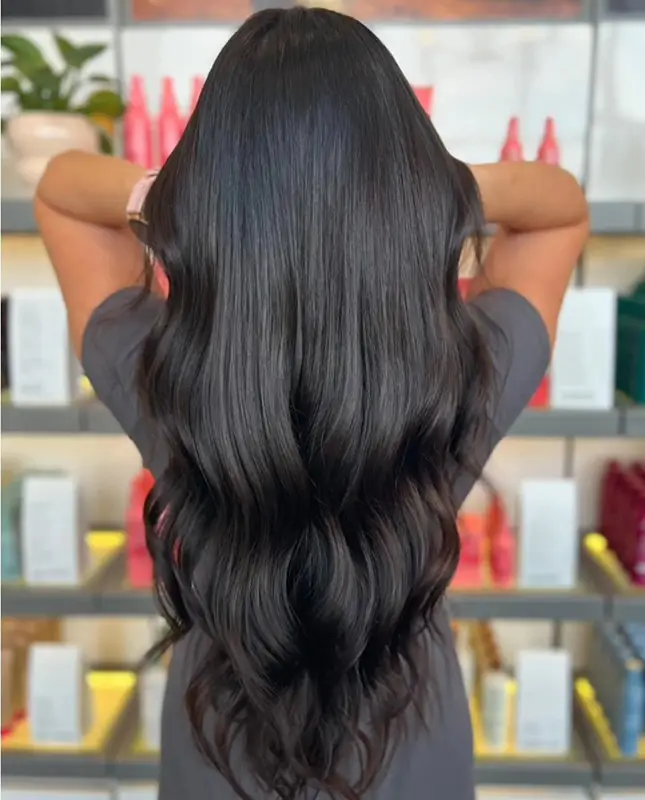 Person with long, wavy, shiny black hair facing away, hands lifted to head, standing in front of shelves with various hair products. - Mango Salon, VA