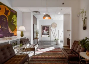 Modern waiting area with brown tufted chairs, a striped rug, artwork on the walls, a large potted plant, and a table lamp; a poster is displayed in the background. - Mango Salon, VA