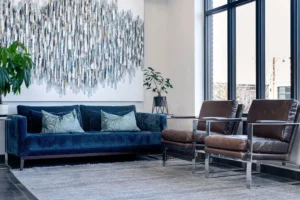 Modern living room with a blue velvet sofa, two brown leather chairs, a large abstract wall art piece, a gray rug, and potted plants by tall windows. - Mango Salon, VA