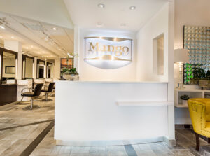 1 Reception area of a modern salon with a white counter and a sign reading "Mango" on the wall; salon chairs and mirrors are visible in the background. - Mango Salon, VA