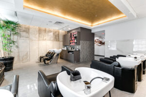 Modern salon interior with black wash stations, white sinks, a display of hair products, a potted plant, and a gold-accented ceiling. The space is bright with minimalist decor. - Mango Salon, VA