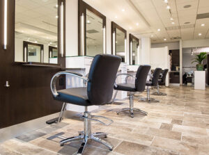 2 Row of empty black salon chairs in front of mirrors and counters in a modern, well-lit hair salon with tiled flooring. - Mango Salon, VA