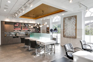 Modern hair salon interior with styling chairs, workstations, product shelves, a chandelier, and large windows letting in natural light. - Mango Salon, VA
