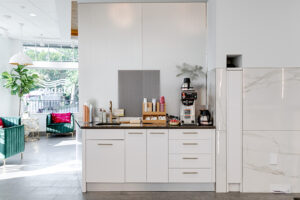 A modern coffee station with white cabinets, a coffee maker, condiments, and cups, situated in a bright, contemporary lounge area with green chairs and large windows. - Mango Salon, VA