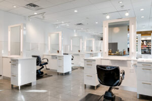 Modern hair salon interior with empty black chairs, white styling stations, large mirrors, and shelves with hair products in the background. Bright lighting and clean, minimalist design. - Mango Salon, VA