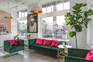 Modern salon waiting area with green velvet sofas, pink cushions, gold tables, indoor plants, and large windows displaying "Margo Salon" signage. - Mango Salon, VA