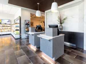8Y6B3577-1 Modern salon reception area with two grey front desks, computer monitors, pendant lights, product shelves, and decorative vase on the counter. - Mango Salon, VA