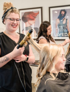 DSC03407 A hairstylist uses a curling iron on a client's hair in a salon, while another stylist works in the background. Both clients are seated and wearing black capes. - Mango Salon, VA