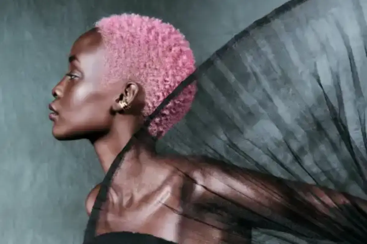 Profile of a person with short pink hair and dark skin, dressed in black clothing with a sheer, pleated black fabric extending behind their shoulder. - Mango Salon, VA