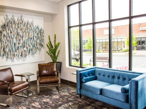 grandopening-mango-18-1 A modern waiting area with two brown leather chairs, a blue tufted sofa, a patterned rug, a large window, abstract wall art, and a tall green plant. - Mango Salon, VA