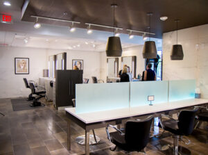 grandopening-mango-23-1 Modern hair salon interior with empty styling chairs, large mirrors, overhead lighting, and a few people near the back. - Mango Salon, VA