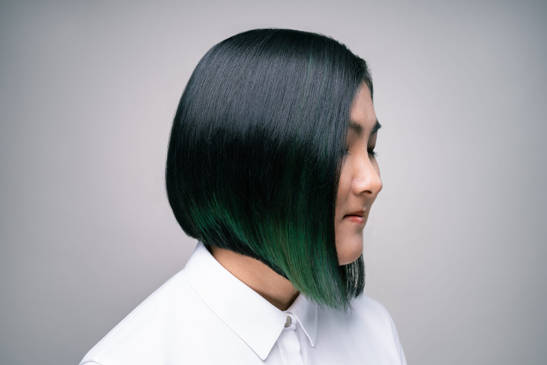 Person with straight, chin-length black hair featuring green tips, wearing a white collared shirt, shown in profile against a plain background. - Mango Salon, VA