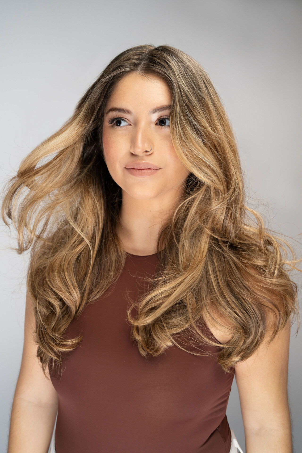 A woman with long, wavy, holiday-ready hair wearing a sleeveless brown top stands against a neutral background, looking slightly to the side. - Mango Salon, VA