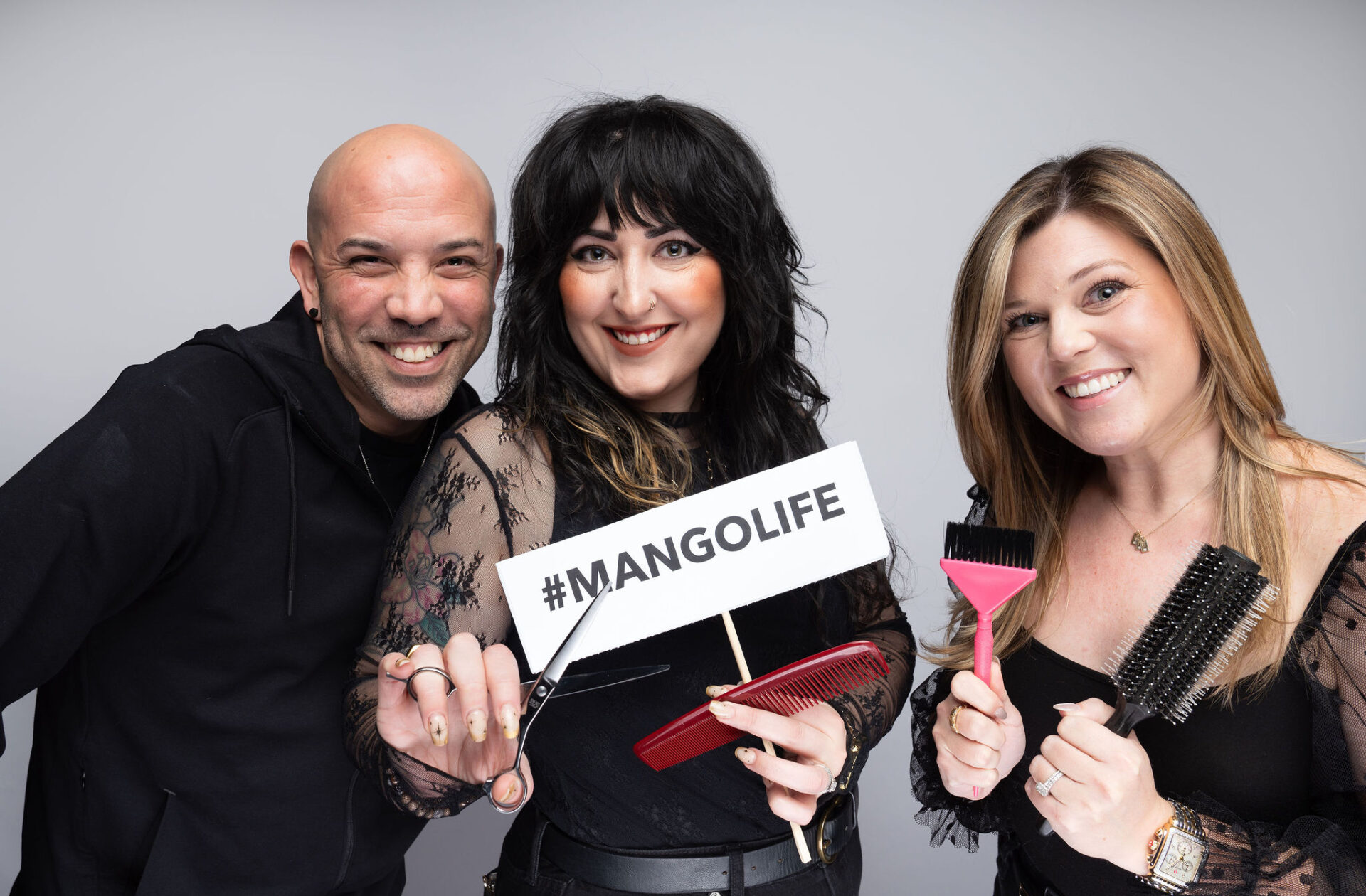 Three people stand smiling, holding hair styling tools and a sign that reads "#MANGOLIFE" against a plain background. - Mango Salon, VA