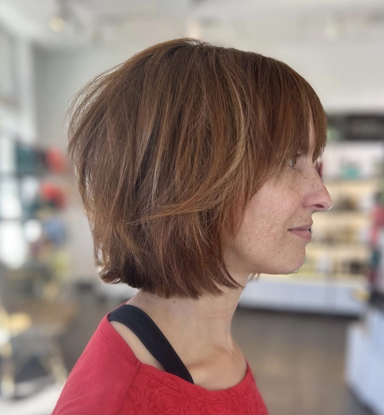 A woman with a chin-length, layered brown bob haircut with subtle highlights, wearing a red top, is shown in profile indoors—a chic nod to bob trends for spring summer hairstyles. - Mango Salon, VA