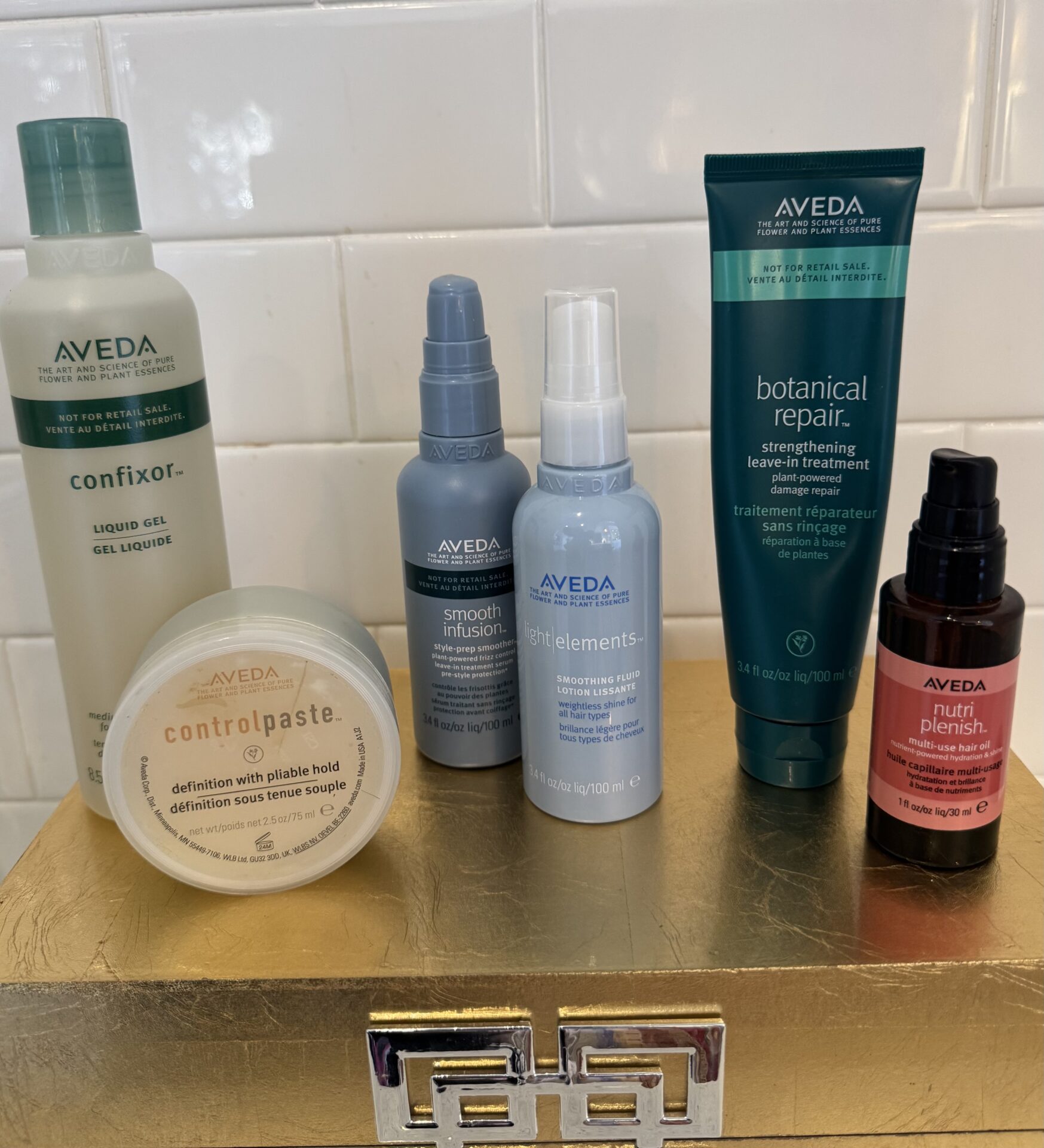 Six Aveda hair styling and treatment products are arranged on a gold box against a white tiled background. - Mango Salon, VA