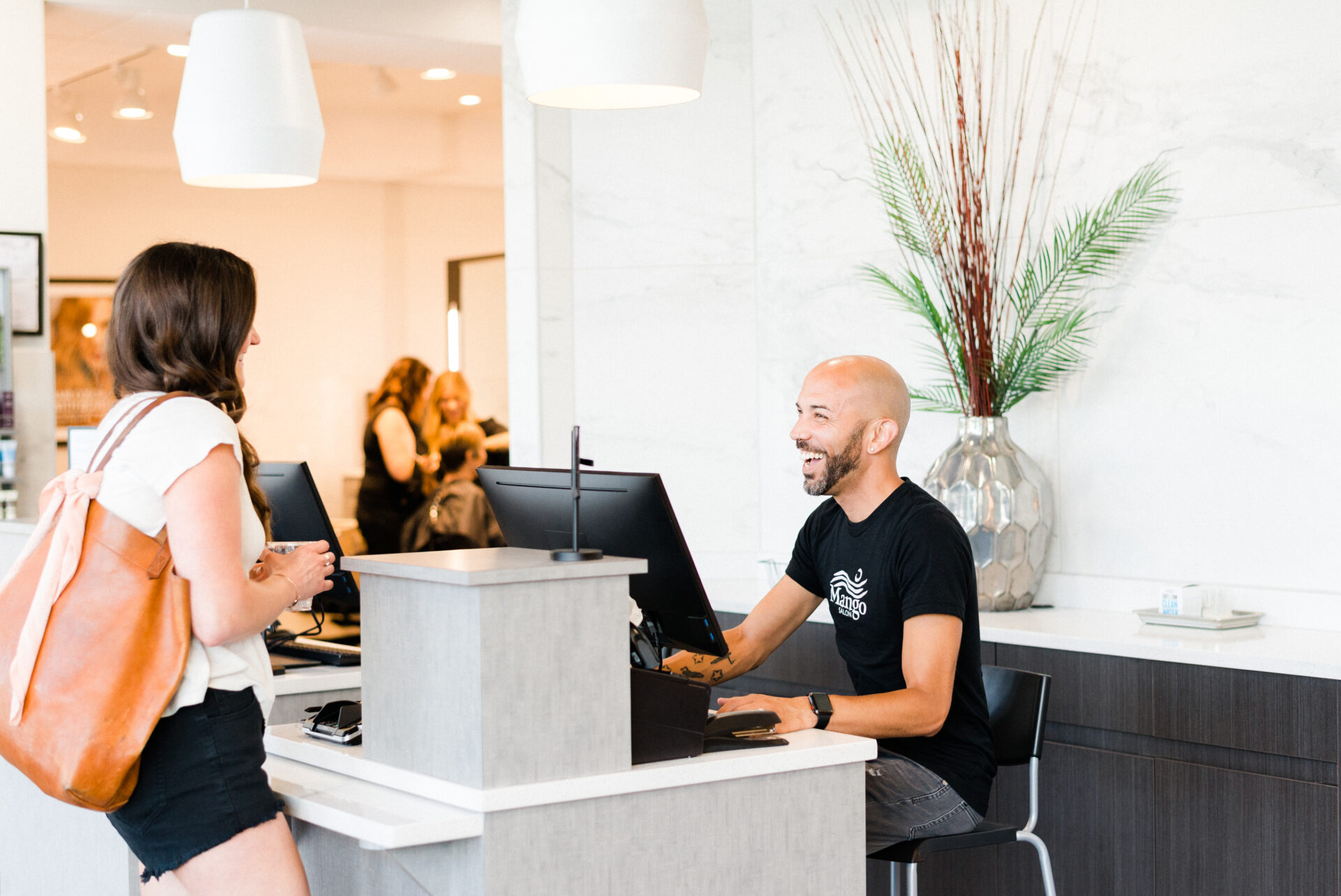 A woman stands at the Mango Salon reception desk speaking to a smiling, seated man—perhaps discussing her leadership career—with a computer monitor and decorative vase in the background. - Mango Salon, VA