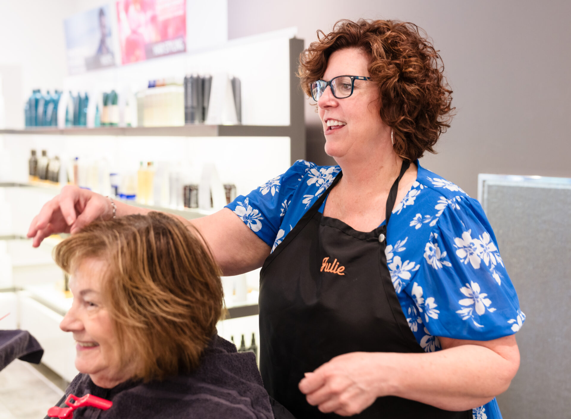 A hairdresser wearing a blue floral shirt and black apron styles a smiling woman's hair in a modern salon. - Mango Salon, VA