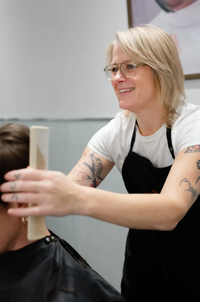 A hairstylist with glasses and tattoos uses a comb while working on a client’s hair in a salon. - Mango Salon, VA