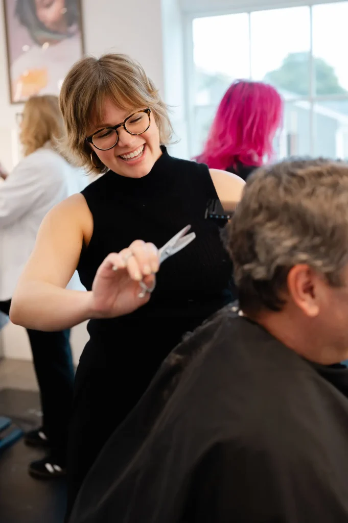 Smiling hairdresser wearing glasses cuts a man’s hair in a bright salon, with another hairstylist and a client in the background. - Mango Salon, VA