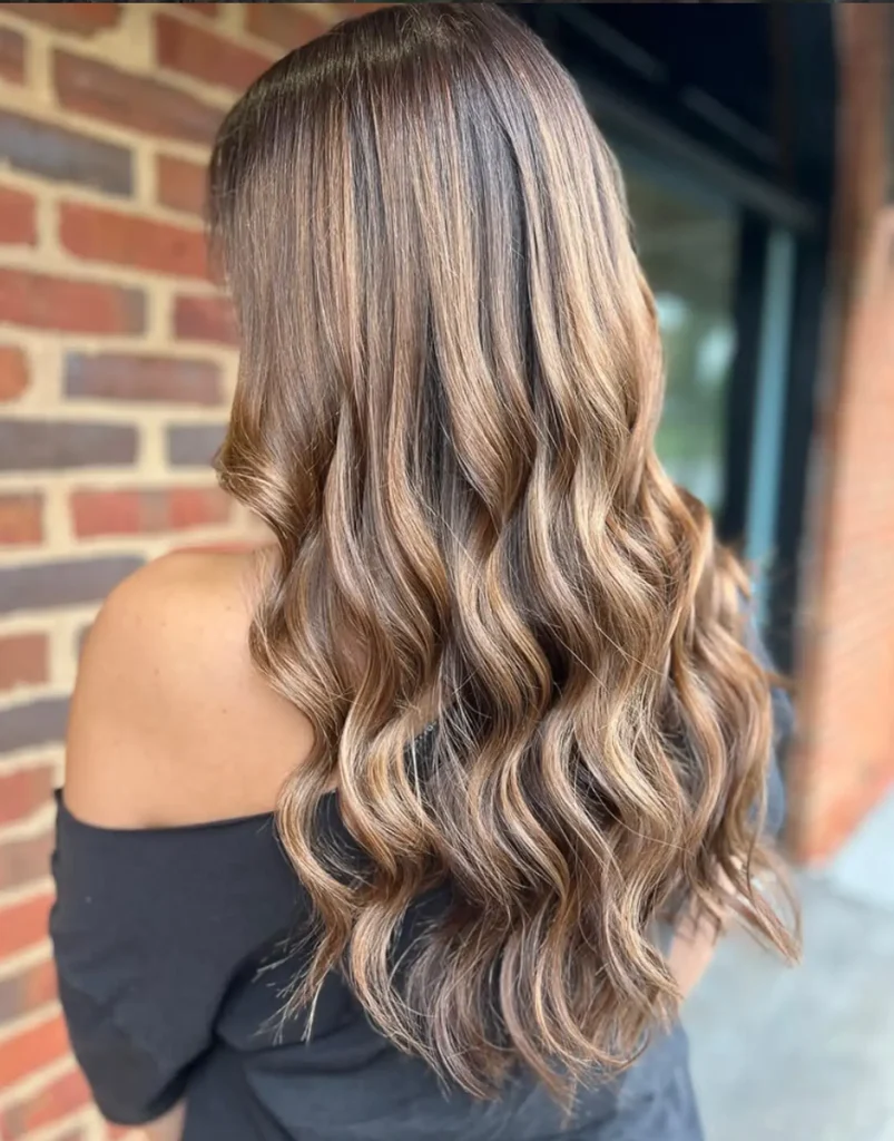 Person with long, wavy, light brown hair with highlights, wearing an off-the-shoulder black top, standing in front of a brick wall. - Mango Salon, VA