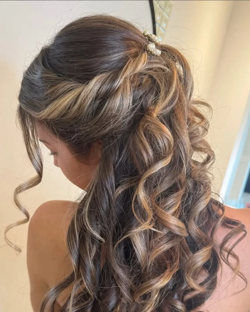 A woman with long, curled brown hair featuring blonde highlights is seen from behind. Her hair is partially pinned up with a decorative hair clip. - Mango Salon, VA