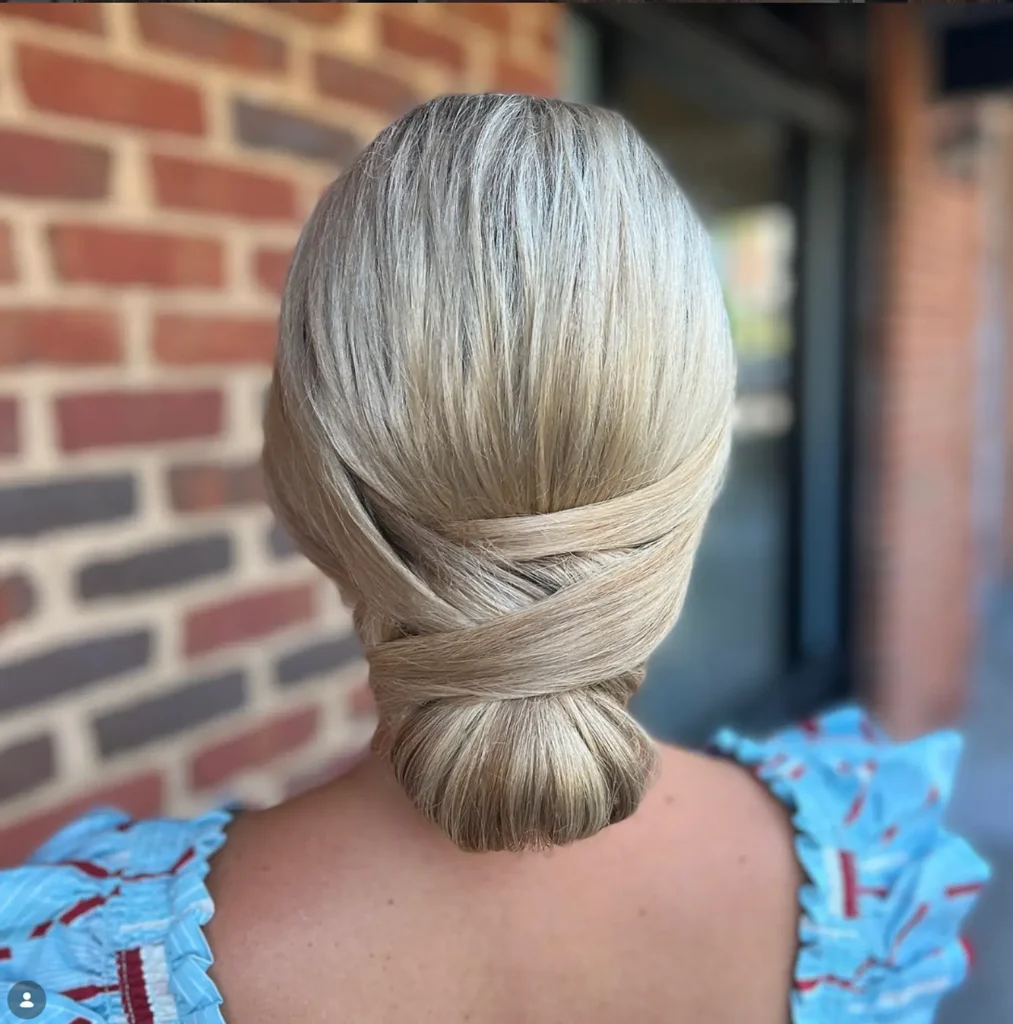 Person with blonde hair styled in a smooth, low twisted chignon bun, standing outdoors in front of a brick wall. - Mango Salon, VA
