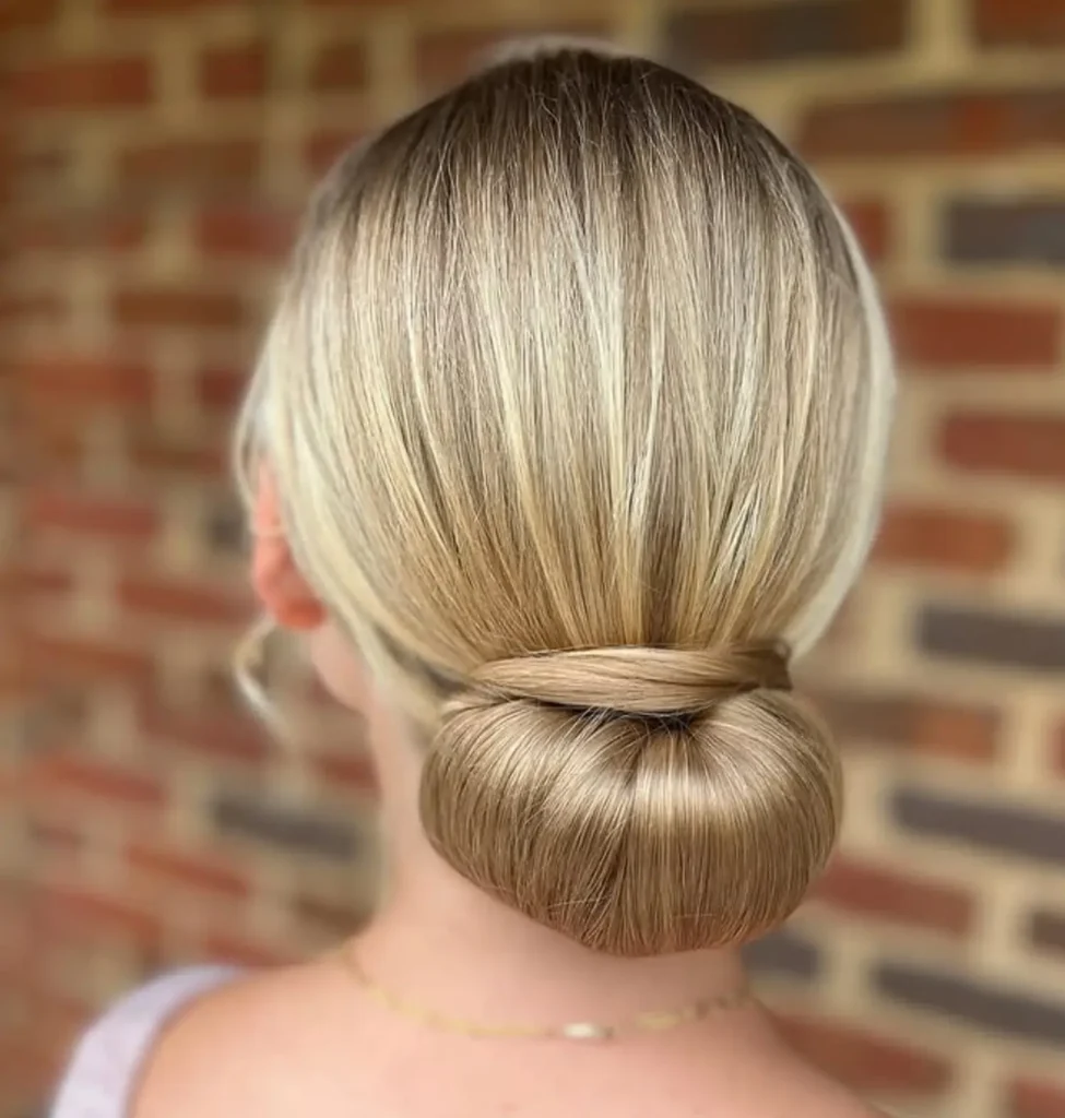 A person with blonde hair styled in a smooth low bun stands in front of a brick wall, facing away from the camera. - Mango Salon, VA