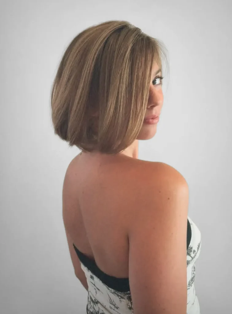 Woman with short, straight, light brown hair in a bob haircut, wearing a strapless top, poses looking over her shoulder against a plain light background. - Mango Salon, VA