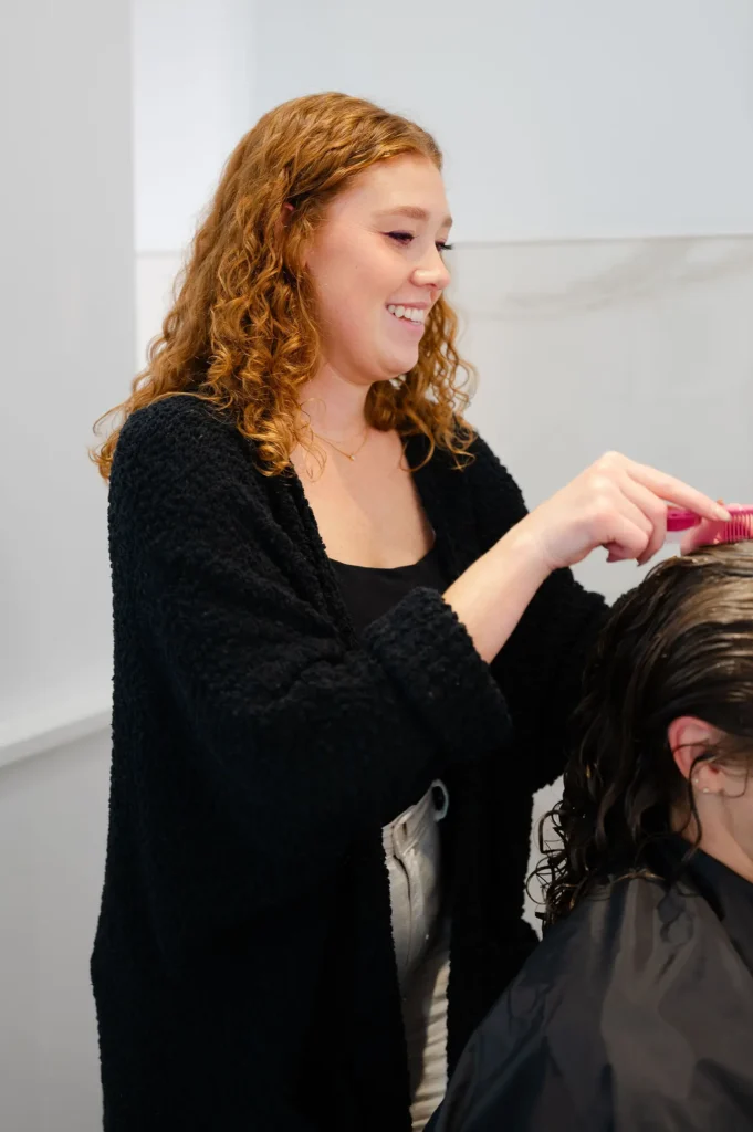 A person with curly hair smiles while combing the wet hair of another person seated in a salon. - Mango Salon, VA