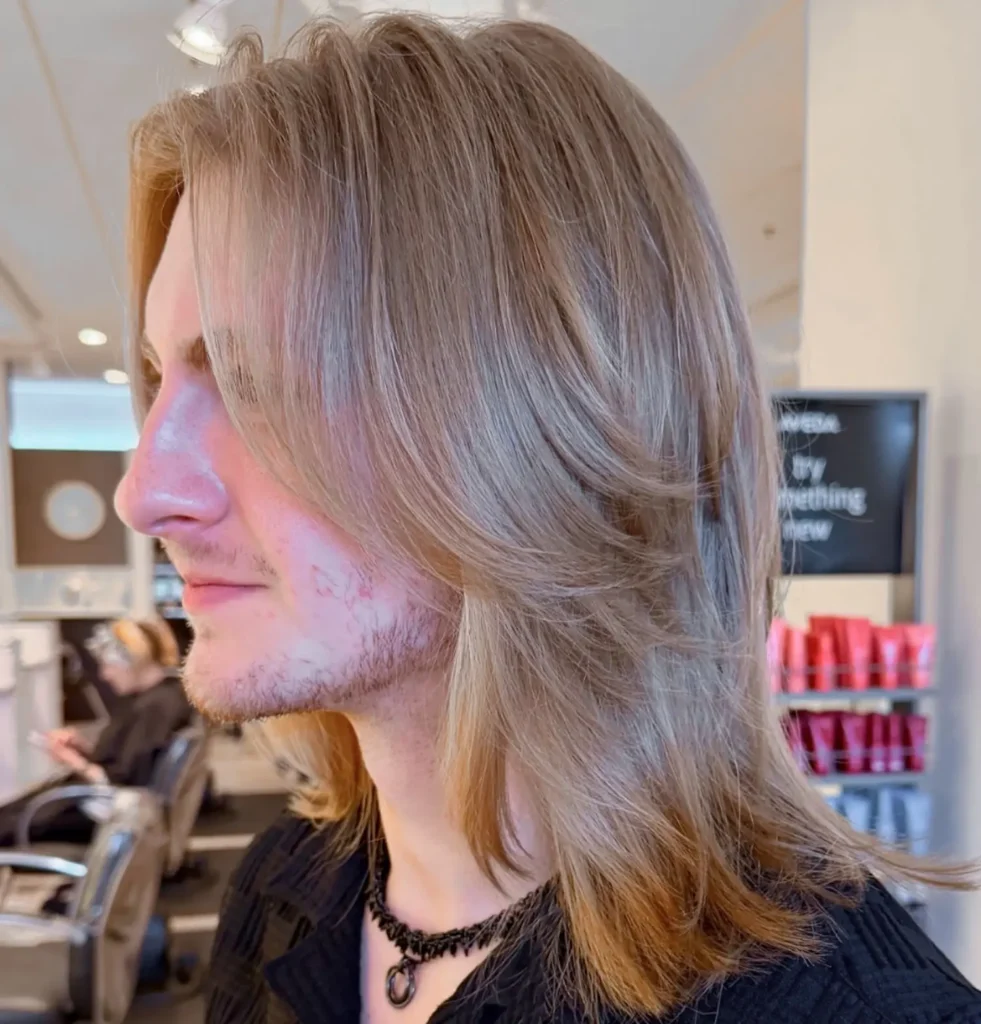 A person with shoulder-length, layered blonde hair and light facial hair is sitting in a salon. Shelves with red hair products are visible in the background. - Mango Salon, VA