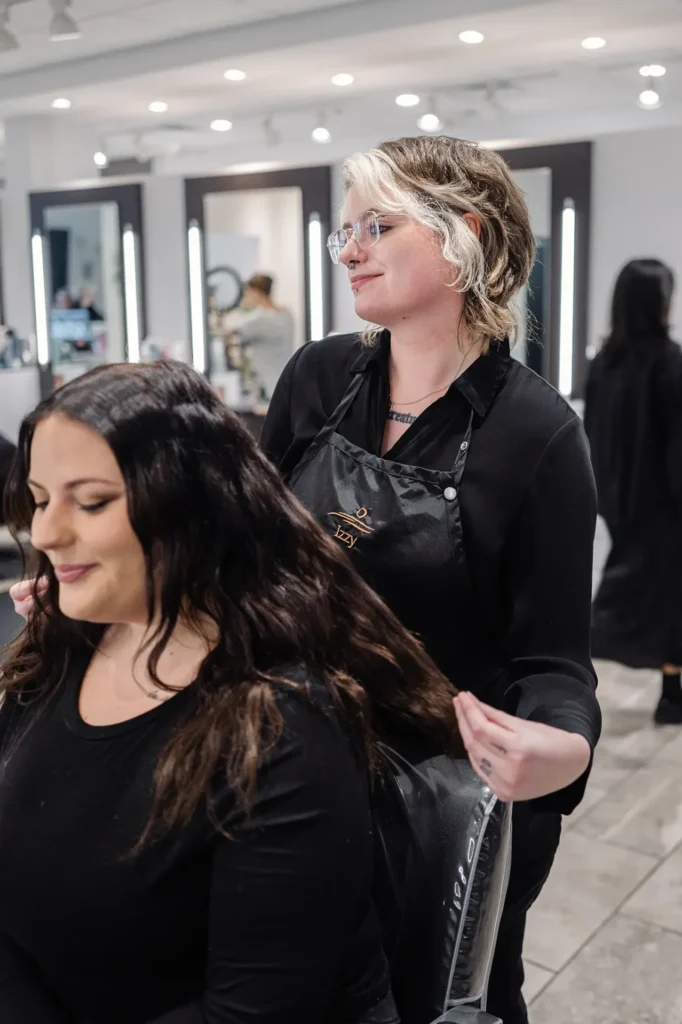 A hairstylist wearing glasses and a black apron is holding a client’s long dark hair in a modern salon with mirrors and lights in the background. - Mango Salon, VA