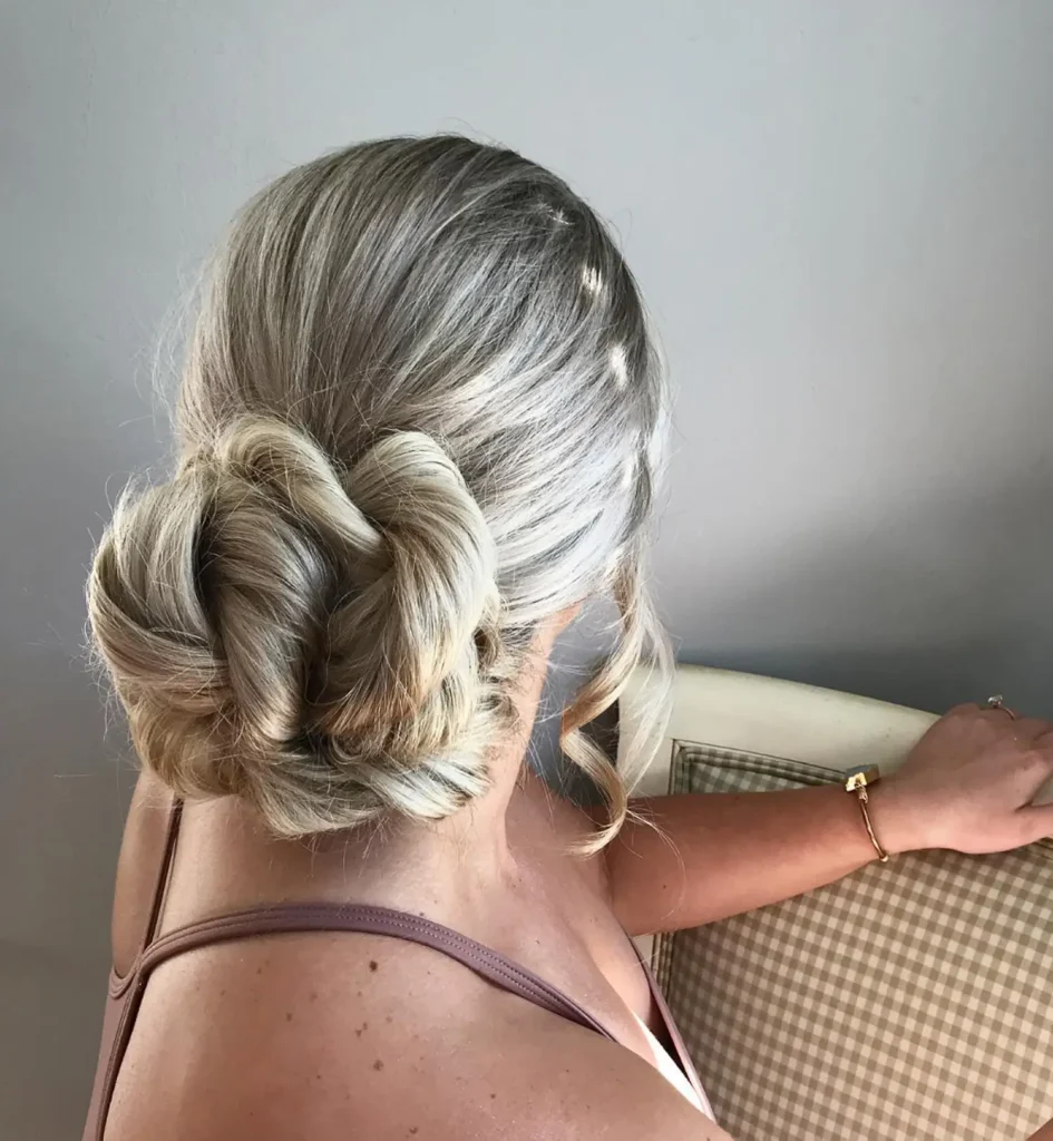 A woman with platinum blonde hair styled in a low twisted bun sits on a checkered chair, with one arm resting on the chairback. - Mango Salon, VA