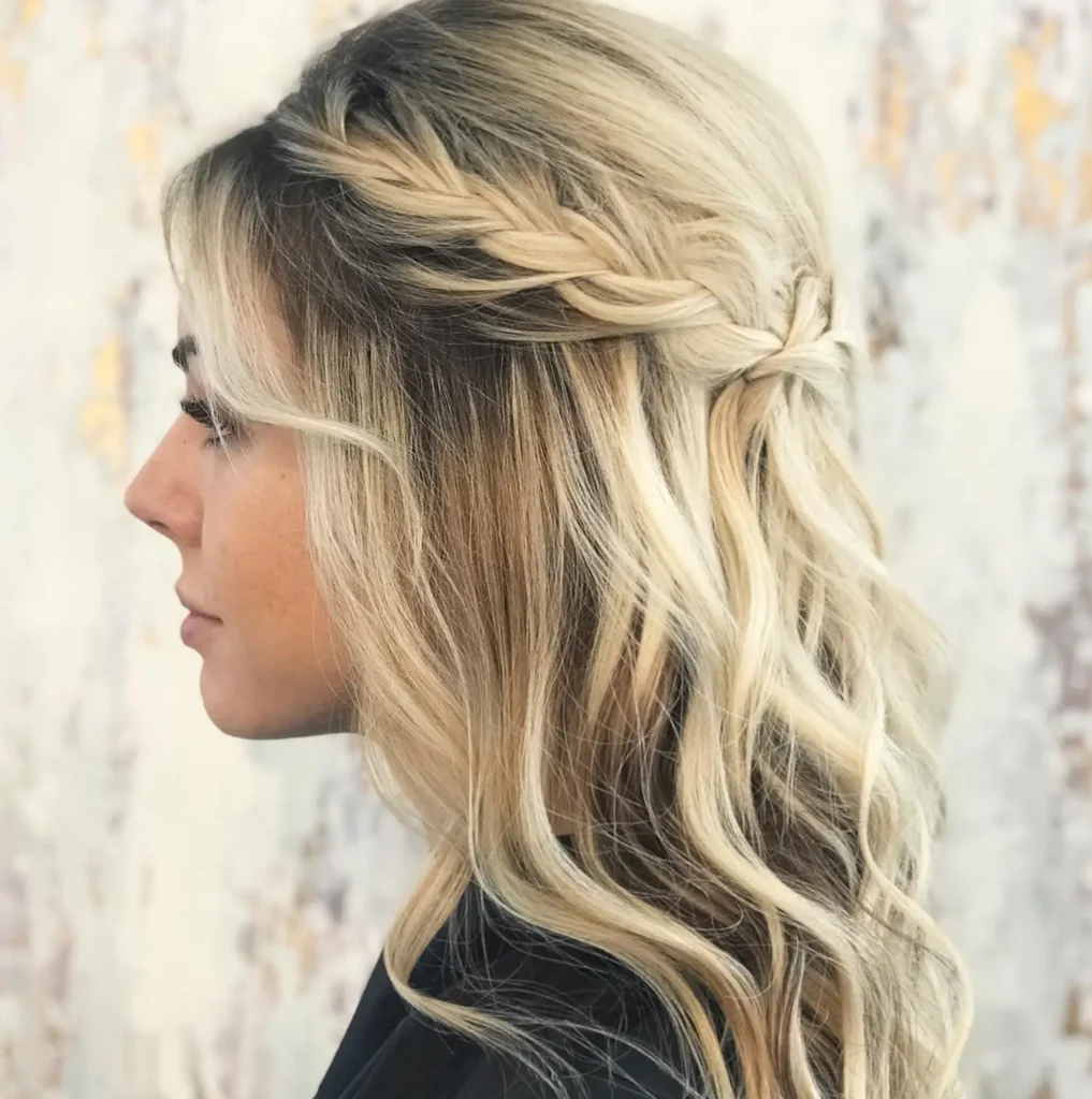 Woman with long, wavy blonde hair styled in loose curls and a side braid, facing left, against a light, blurred background. - Mango Salon, VA