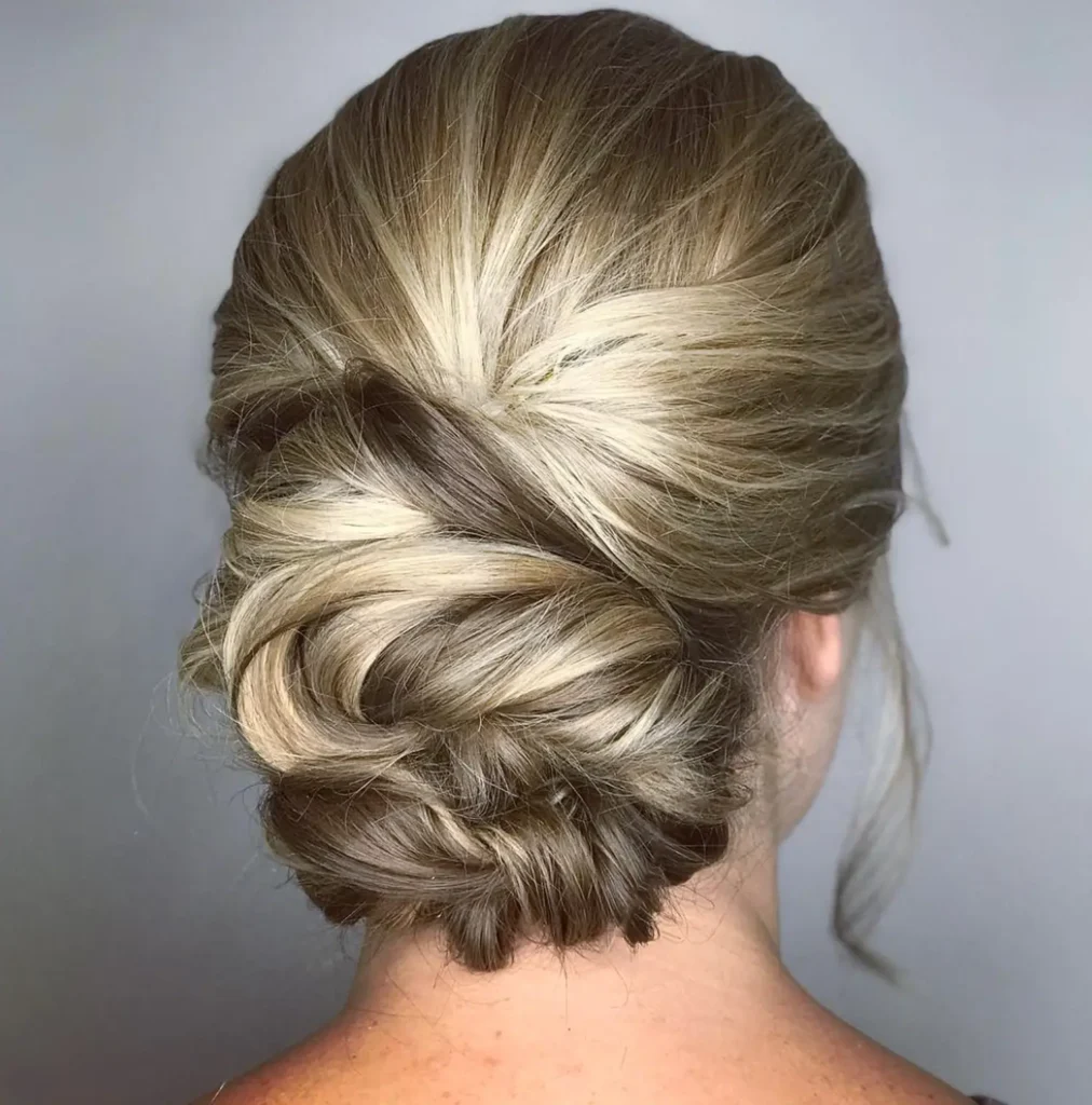 A woman with blonde and brown hair styled in a low, textured updo with a loose, twisted bun, photographed from behind against a plain background. - Mango Salon, VA