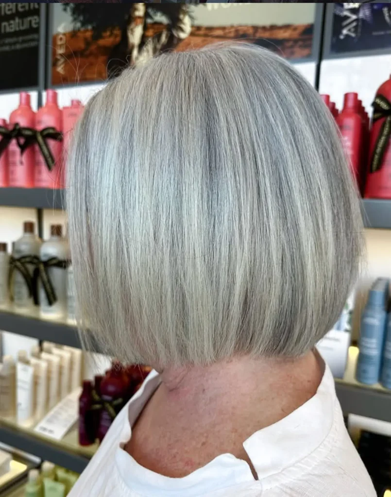 Person with straight, chin-length gray hair in a blunt bob haircut, viewed from the side in a salon setting with hair products on shelves in the background. - Mango Salon, VA