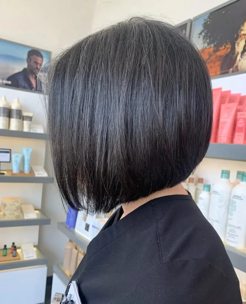 Person with straight, chin-length black bob haircut facing sideways in a salon setting with shelves of hair products in the background. - Mango Salon, VA