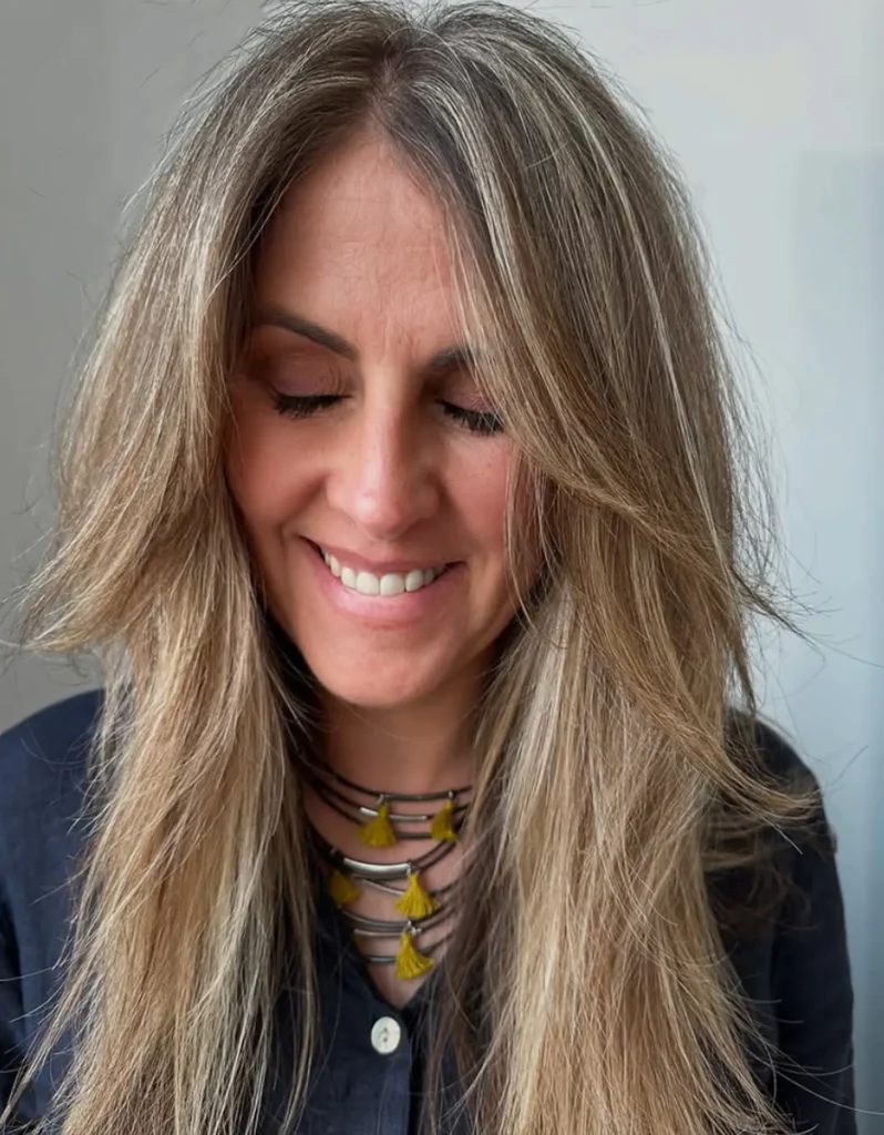Woman with long, layered, highlighted hair smiles with eyes closed, wearing a dark shirt and a necklace with yellow tassels, against a plain background. - Mango Salon, VA