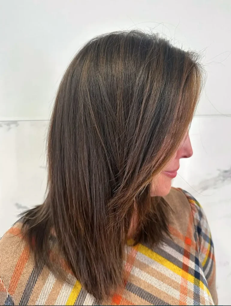 Woman with straight, shoulder-length brown hair featuring subtle highlights, facing right. She is wearing a beige plaid-patterned top. The background is light with a faint marble texture. - Mango Salon, VA