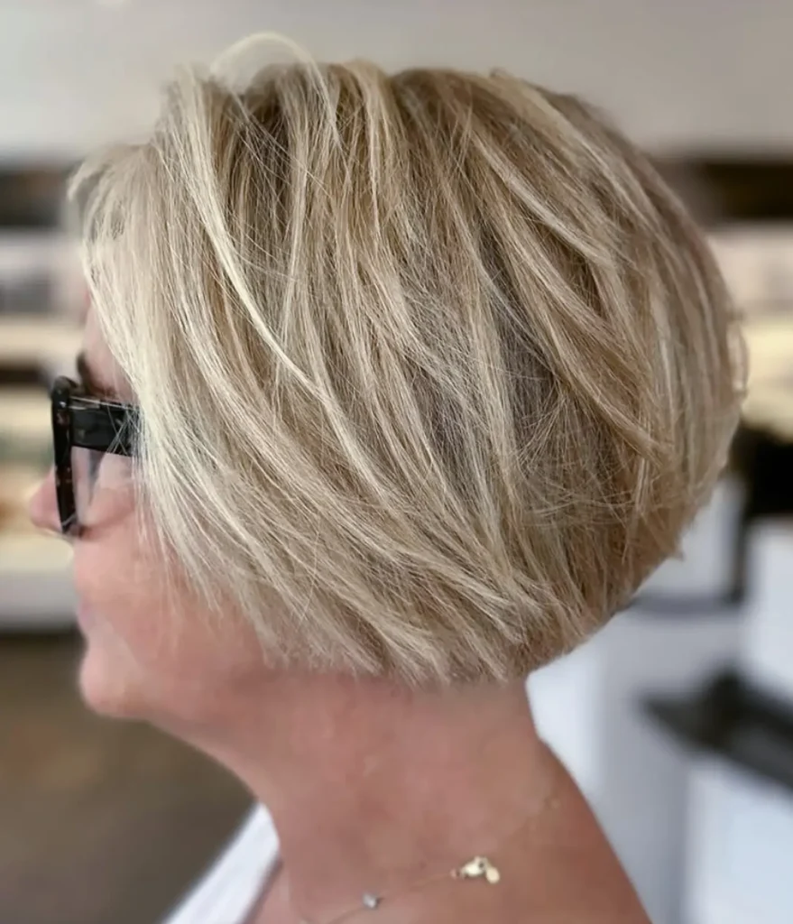 A person with short, layered blonde hair in a bob cut, wearing black glasses and a gold necklace, shown from the side. - Mango Salon, VA