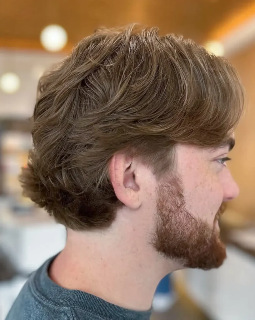 A young man with light brown wavy hair and a trimmed beard is shown in profile, indoors with a blurred background. - Mango Salon, VA