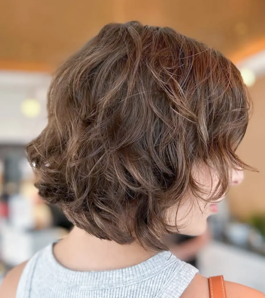 Person with short, wavy brown hair wearing a light gray sleeveless top, shown from the back and side in an indoor setting. - Mango Salon, VA