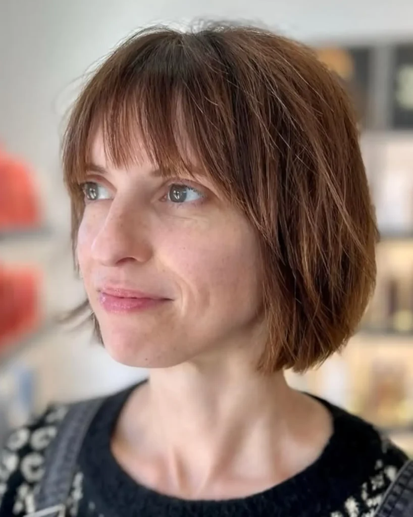 A person with short brown hair and bangs looks slightly to the left, wearing a black and white sweater. The background is softly blurred with some books or artwork visible. - Mango Salon, VA