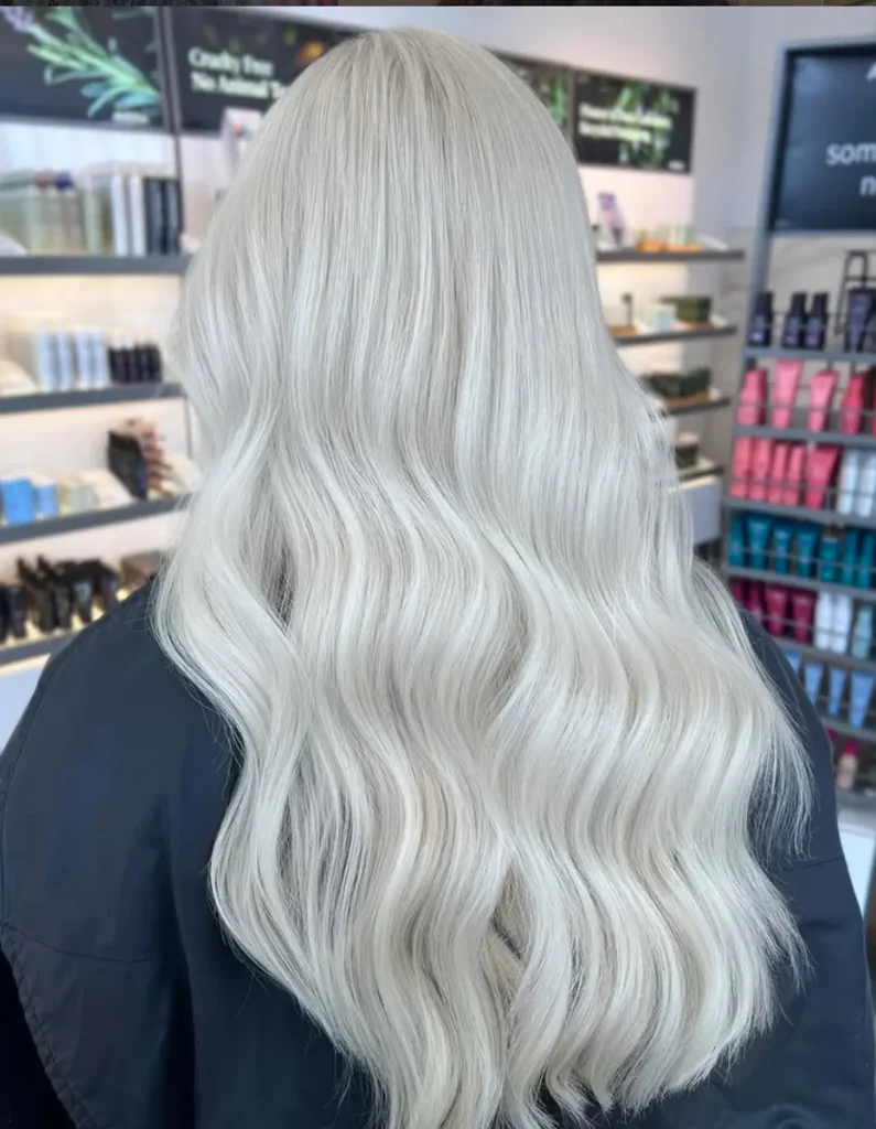 Person with long, wavy, platinum blonde hair seen from behind, standing in a salon with shelves displaying various hair products. - Mango Salon, VA